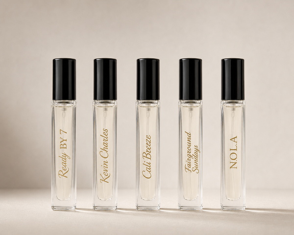 Five clear glass rollerball perfume bottles with black caps and gold script labels, arranged in a row on a warm beige background. The labels read: Ready BY 7, Kevin Charles, Cali Breeze, Fairground Sundays, and NOLA.