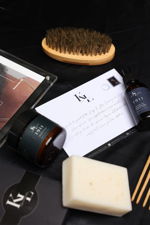 A flat-lay view of a grooming or skincare set on a black background, including a cream-colored soap bar, a dark amber jar, a white handwritten card, a small dark bottle, wooden chopsticks, a wooden-handled brush, and a black box with a stylized K logo.