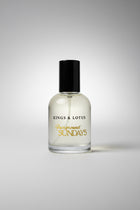 A clear glass bottle of Kings & Lotus 'Fairground Sundays' fragrance with a black cap, resting on a black textured surface with a branded card in the background.