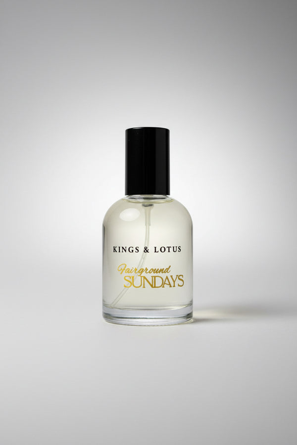 A clear glass bottle of Kings & Lotus 'Fairground Sundays' fragrance with a black cap, resting on a black textured surface with a branded card in the background.