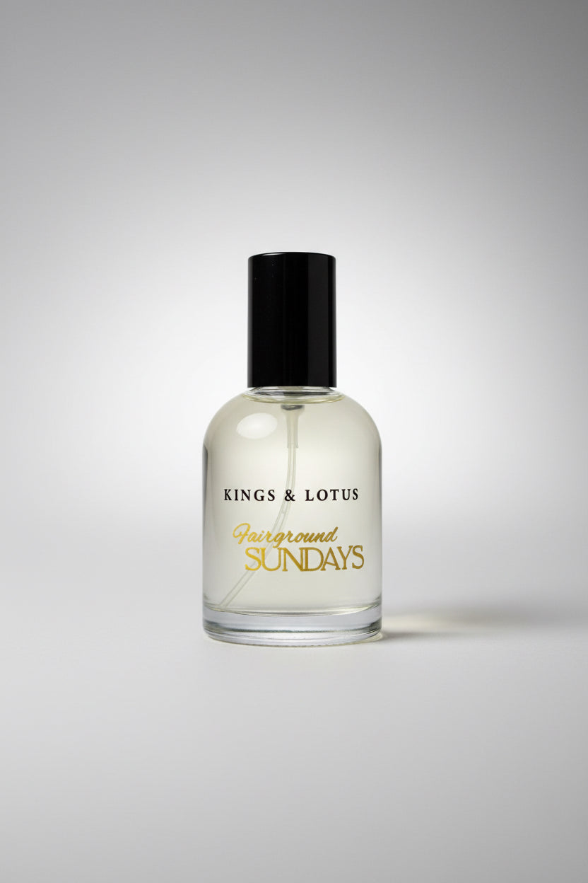 A clear glass bottle of Kings & Lotus 'Fairground Sundays' fragrance with a black cap, resting on a black textured surface with a branded card in the background.