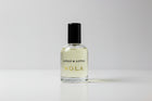 A bottle of Nola grooming essence and lotus by Kings and Lotus, featuring a clear glass bottle with a black cap and gold lettering, resting on a black fabric surface.