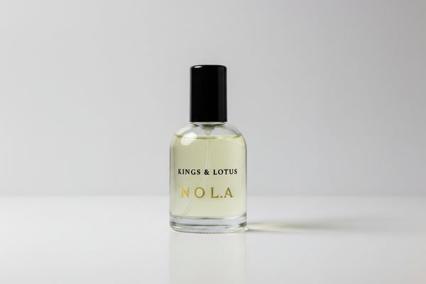 A bottle of Nola grooming essence and lotus by Kings and Lotus, featuring a clear glass bottle with a black cap and gold lettering, resting on a black fabric surface.