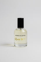 A bottle of Kings & Lotus Ready By 7 fragrance, featuring a clear glass bottle filled with light yellow liquid and a black cap, resting on a black surface with a branded black box in the background.