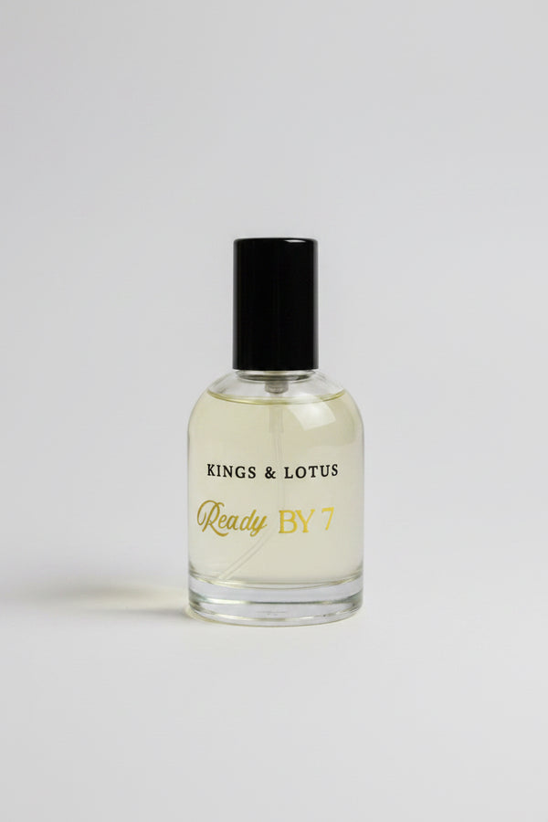A bottle of Kings & Lotus Ready By 7 fragrance, featuring a clear glass bottle filled with light yellow liquid and a black cap, resting on a black surface with a branded black box in the background.