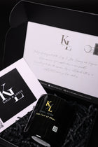 A black cylindrical bottle with gold lettering sits inside a black gift box with shredded paper. A white card with a black logo and a handwritten letter are arranged around it.