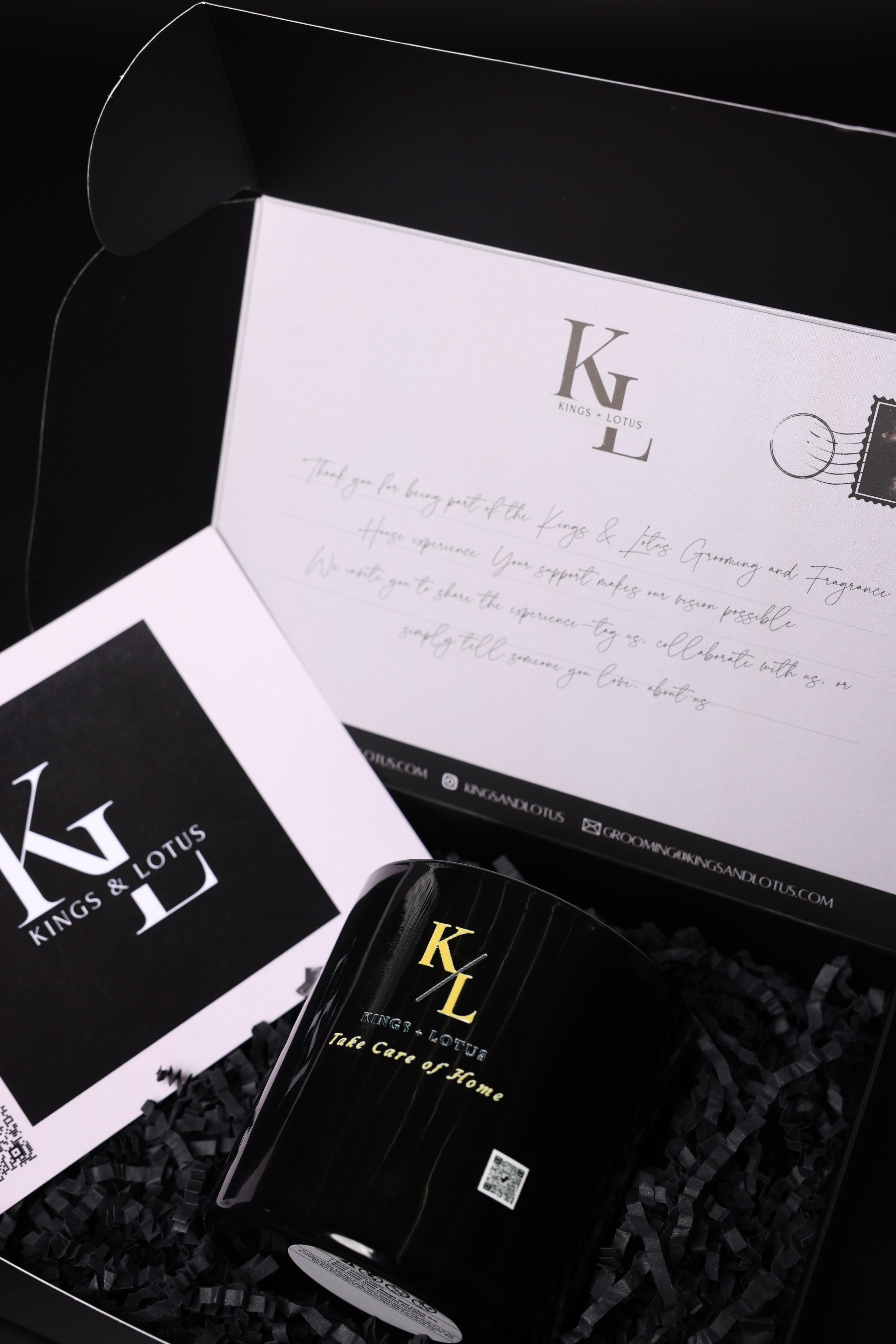 A black cylindrical bottle with gold lettering sits inside a black gift box with shredded paper. A white card with a black logo and a handwritten letter are arranged around it.
