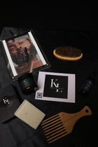 A flat-lay arrangement of hair care and grooming products on a black fabric background, including a wooden wide-tooth comb, a beige shampoo bar, a dark amber bottle, a small black tube, a printed card with a QR code and the K1 logo, a black box with a K1 logo, and a hair brush with a wooden handle.