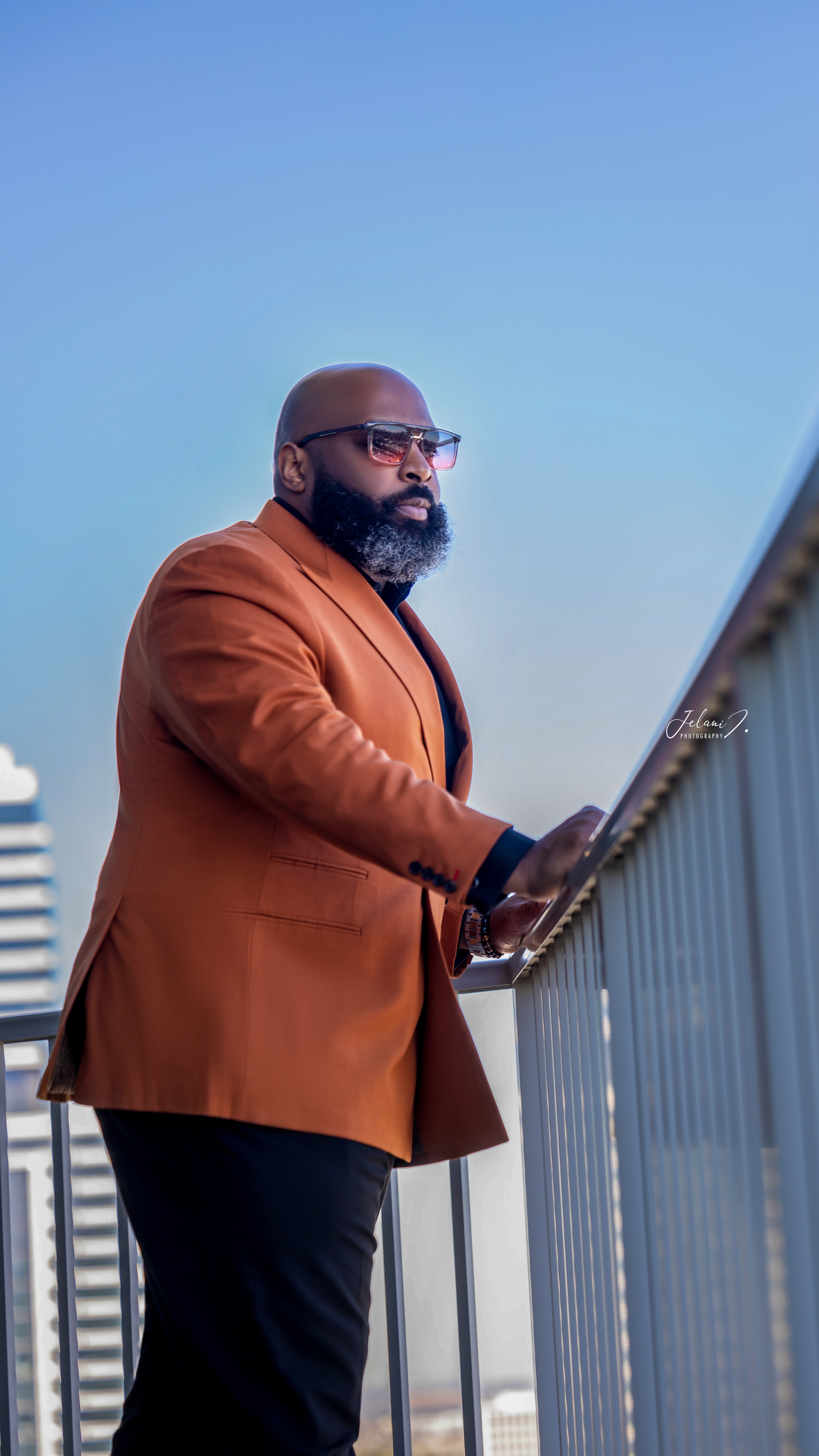 Man in an orange blazer standing on a balcony with a clear blue sky